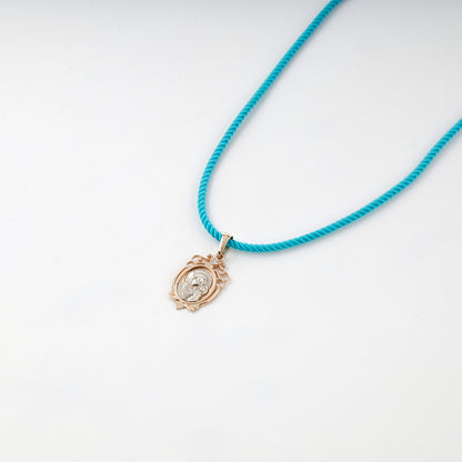 Two-tone gold icon pendant shown on a turquoise styling cord